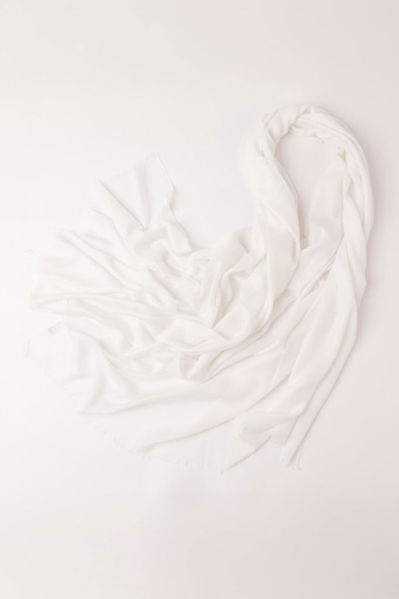 The Ivory Whisper 100% Cashmere Scarf by looselyboho is displayed on a plain, light surface, showcasing its minimalist luxury with a soft texture and frayed edges, some sections loosely folded and others elegantly spread out.