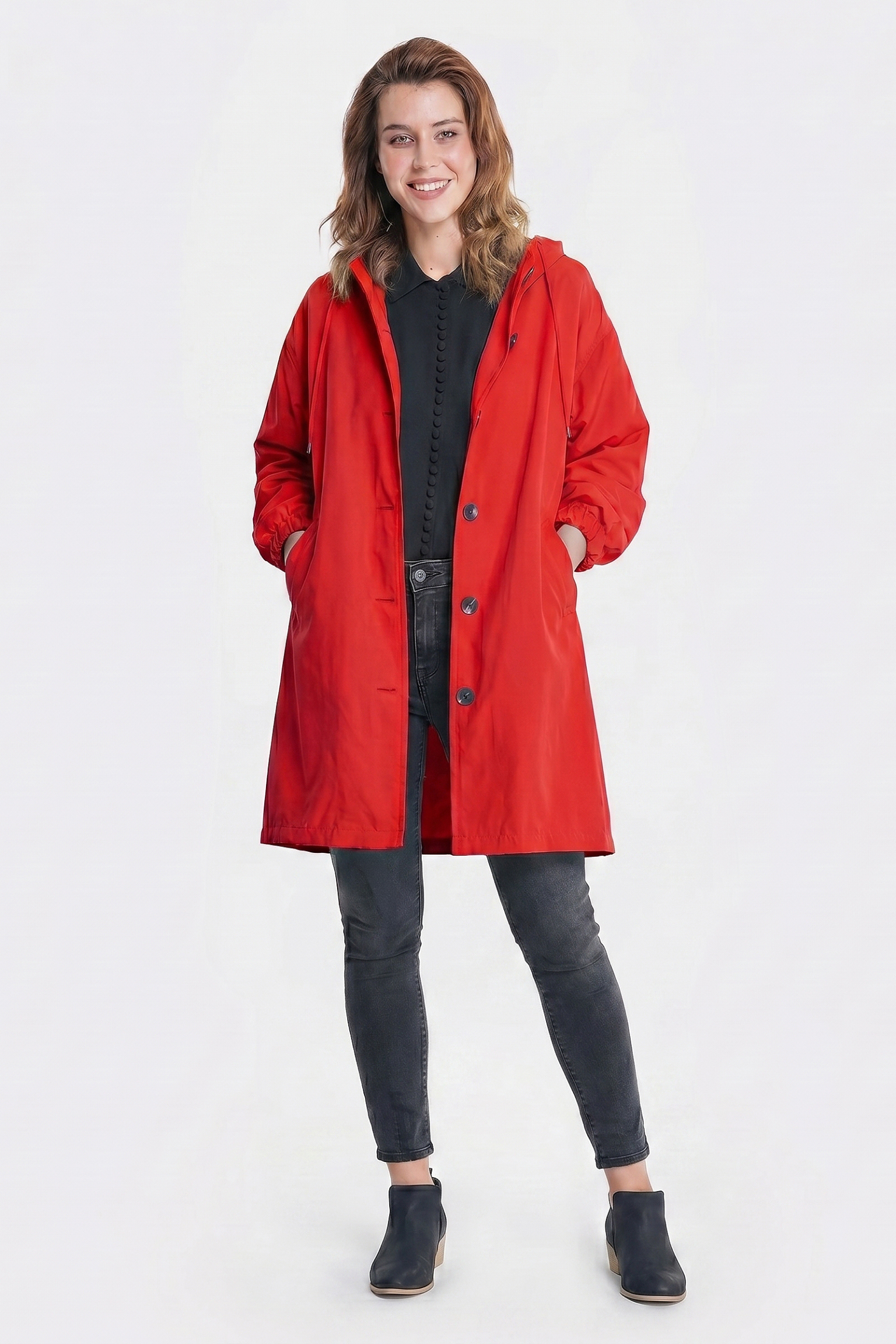 A person stands facing forward, smiling in the Icon Windbreaker Rain Jacket—a long red water-resistant coat—over a black top, dark jeans, and black shoes, with hands in the pockets against a plain white background.