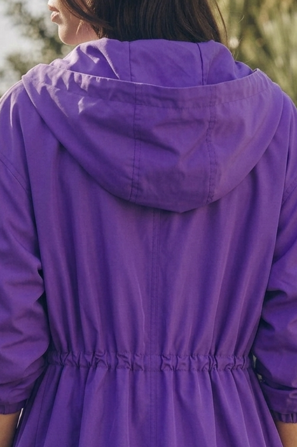 A person with long brown hair is shown from behind, wearing the Icon Rain Windbreaker Jacket in purple—a water-resistant hooded jacket with an elastic waistband. The background features blurred greenery.