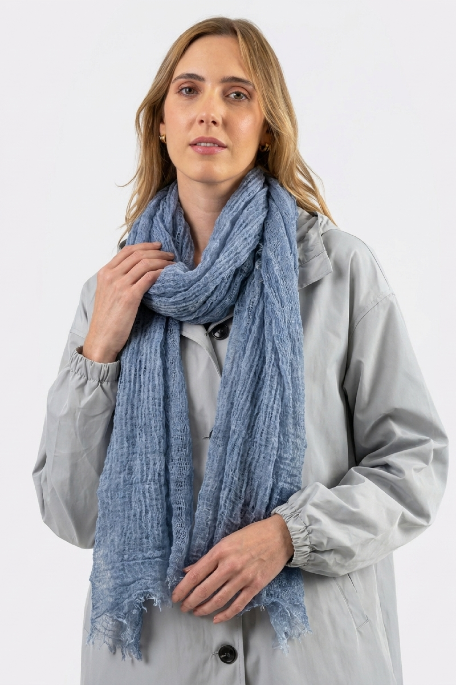 A woman in a light gray coat and the Lightweight Linen Scarf stands against a white background, gazing at the camera with a neutral expression. This versatile accessory subtly enhances her outfit’s texture.
