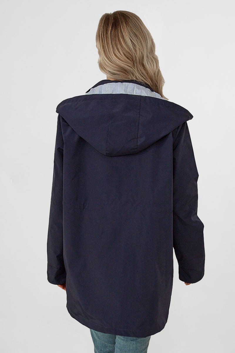 A person with long, wavy hair is shown from the back, wearing a looselyboho STRIPED NAVY SHADOW WINDBREAKER JACKET. The hood is down, revealing a blue and white striped lining inside. The individual is also wearing light blue jeans. The background is plain white.