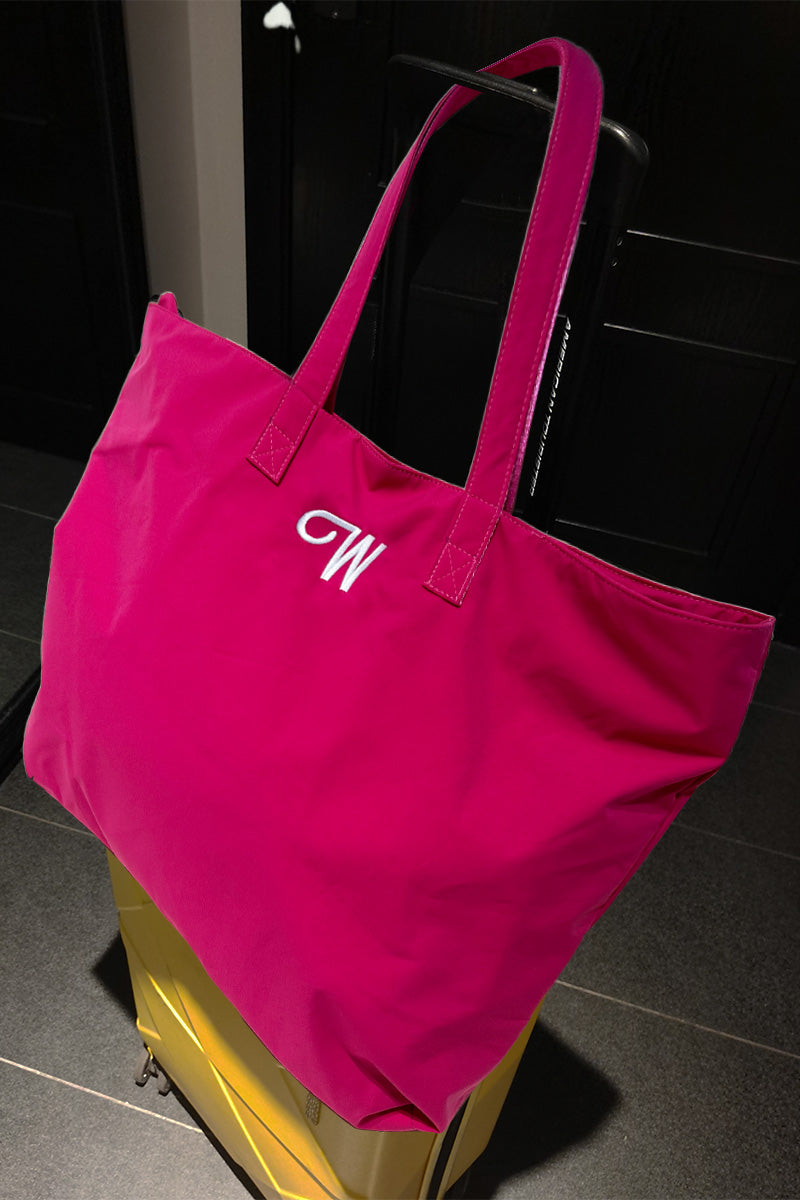A bright "The One" Barbara Pink Waterproof Travel Bag by looselyboho, featuring a white embroidered letter "W", is placed on top of a yellow suitcase. The setting appears to be indoors, possibly a hallway or a room.