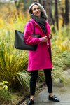 A person with long gray hair stands outdoors on a path surrounded by tall grass and trees. They are wearing a bright pink coat, black pants, and black shoes. The coat is the BARBARA PINK WINDBREAKER OUTFIT from looselyboho's limited edition collection and perfectly echoes that iconic Barbie Pink hue. They carry a large black handbag over one shoulder and have a small smile on their face.