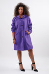 Against a white backdrop, a person confidently wears The Windbreaker Jacket's GINGHAM ULTRAVIOLET WINDBREAKER, its large buttons and pockets accentuating the garment. The ultraviolet hue highlights their curly hair and black high-heeled shoes as they face forward with one hand in a pocket.