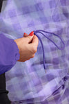 A hand with red nail polish holds an ultraviolet drawstring against a backdrop of purple and white checkered fabric, while wearing The Windbreaker Jacket's GINGHAM ULTRAVIOLET WINDBREAKER.