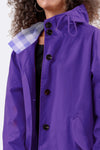 A person with curly hair and hoop earrings wears The Windbreaker Jacket's GINGHAM ULTRAVIOLET raincoat, showcasing its checked collar. It's water-resistant with large buttons and a drawstring hood, set against a plain white background.