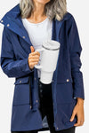 A person wearing a blue jacket, white shirt, and black pants is holding a large white looselyboho Indestructible Travel Cup with a handle. The jacket has multiple buttons and pockets, and the person’s hair appears gray and wavy. The background is plain white.