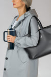 A person with grey hair is wearing a light grey raincoat and carrying a large grey tote bag over their shoulder. In their left hand, they hold a dark grey, spill-proof looselyboho Indestructible Travel Cup with a blue lid and straw. The background is plain and neutral.