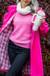 A person with long gray hair is dressed in a bright pink checkered coat, pink sweater, and dark pants. They wear a white scarf and hold a large "Indestructible Travel Cup" by looselyboho. The background has dark foliage. The person's face is not visible.