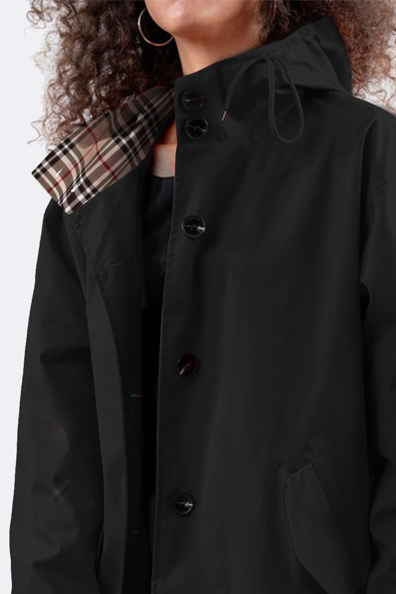 A person with curly hair is wearing a looselyboho LUXE BLACK WATERPROOF WINDBREAKER featuring large buttons and an adjustable hood lined with plaid. The person's face is hidden, adding mystery to their intriguing appearance.