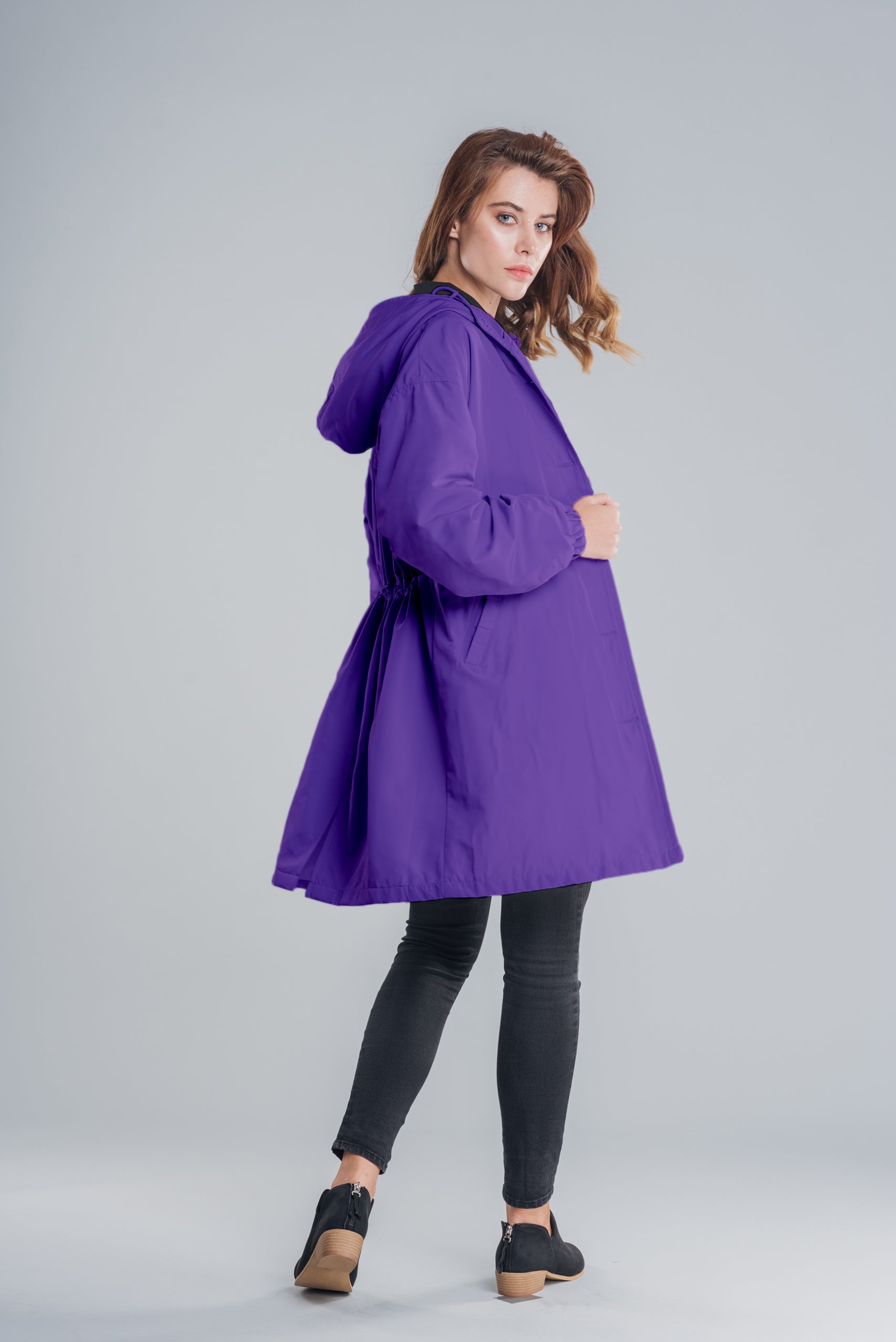 A woman is standing and facing slightly to the side, showcasing the versatility of her purple Water Resistant Oversized Hooded Windbreaker Rain Jacket by The WJacket, paired with black leggings and black shoes. The plain light gray background highlights her light brown, wavy hair.