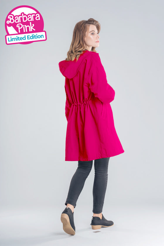 A woman stands looking over her shoulder, wearing a bright pink Water Resistant Oversized Hooded Windbreaker Rain Jacket from The WJacket. She is also dressed in dark pants and black shoes. Text on the image reads: "Barbara Pink Limited Edition." The outfit exudes timeless style and versatility.
