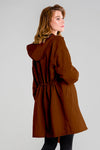 A person with long hair stands facing away, wearing the looselyboho BURNT BROWN WINDBREAKER. This dark brown hooded coat is crafted from lightweight, water-resistant fabric and features gathered detailing at the back. Its adjustable hood frames their side-turned gaze against a plain light gray background.