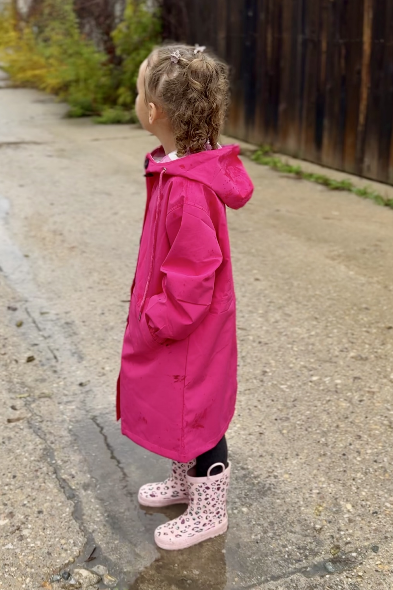 A young child with curly hair stands on a wet outdoor surface, wearing the looselyboho Bright Berry Kids’ Windbreaker and pink patterned rain boots, looking to the side. A wooden fence and greenery are visible in the background.
