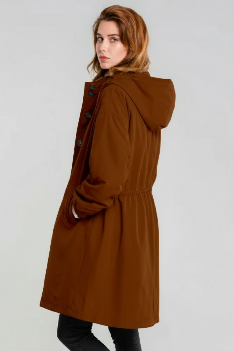 A person with light skin and auburn hair models the looselyboho BURNT BROWN WINDBREAKER, featuring an adjustable hood and buttons, with one hand in a pocket, standing against a plain light background.
