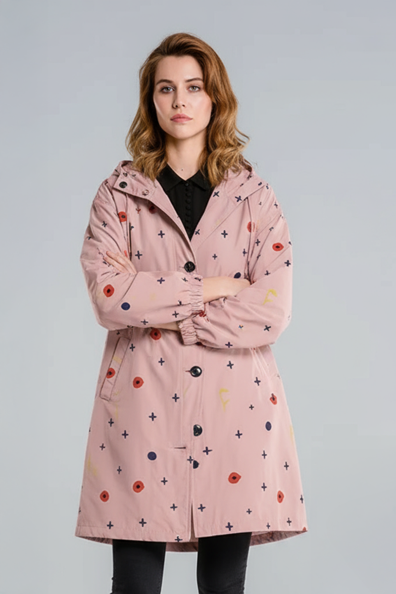 A woman stands with her arms crossed, wearing the looselyboho DOT & BLOOM WINDBREAKER—a light pink, water-resistant jacket with a button front and small navy, red, and yellow geometric shapes—paired with black pants against a plain gray background.