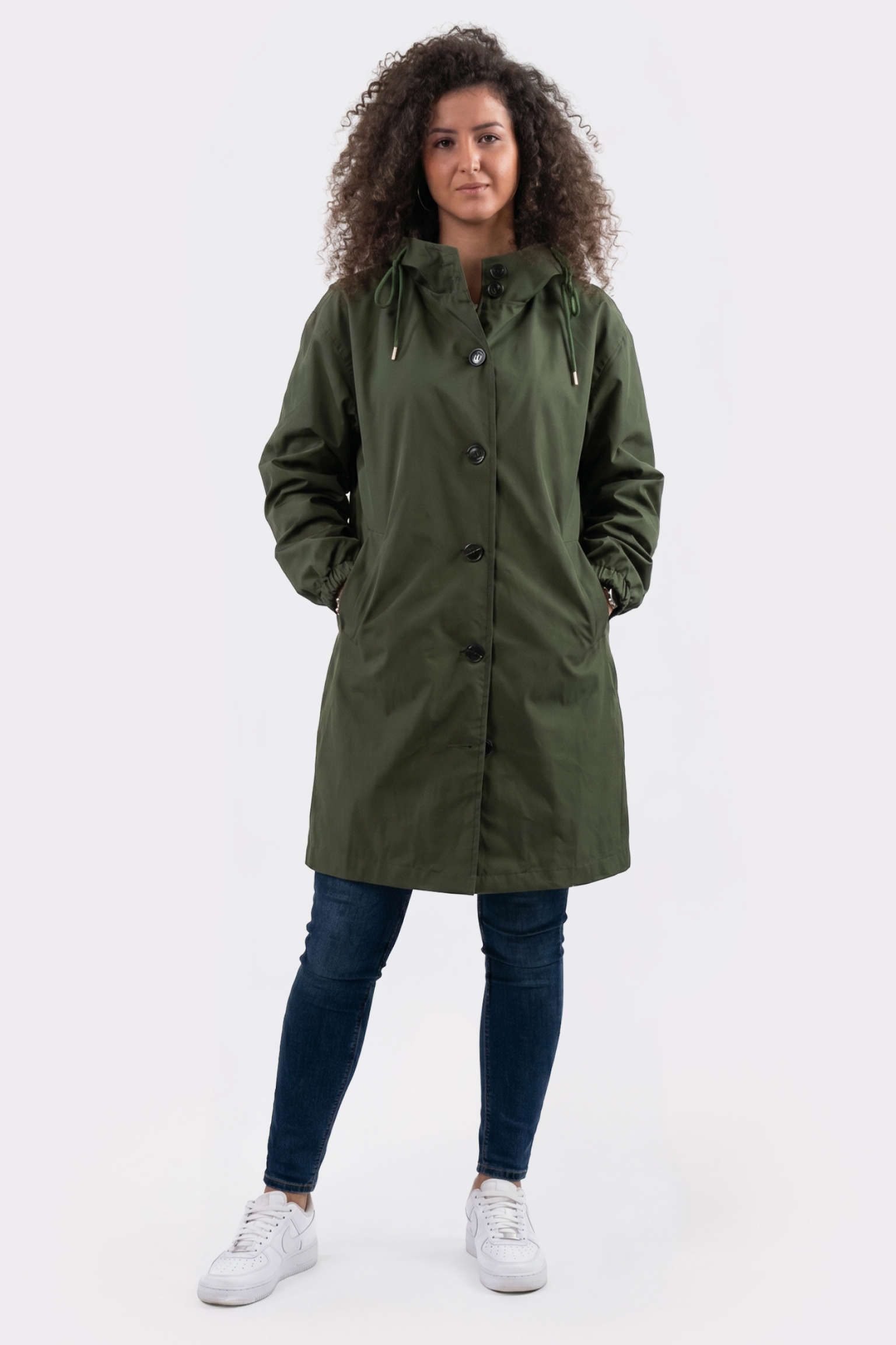 A person with curly hair wears the Icon Windbreaker Rain Jacket, styled in a long olive green design, along with blue jeans and white sneakers. They stand facing forward, hands in pockets, against a plain white background.