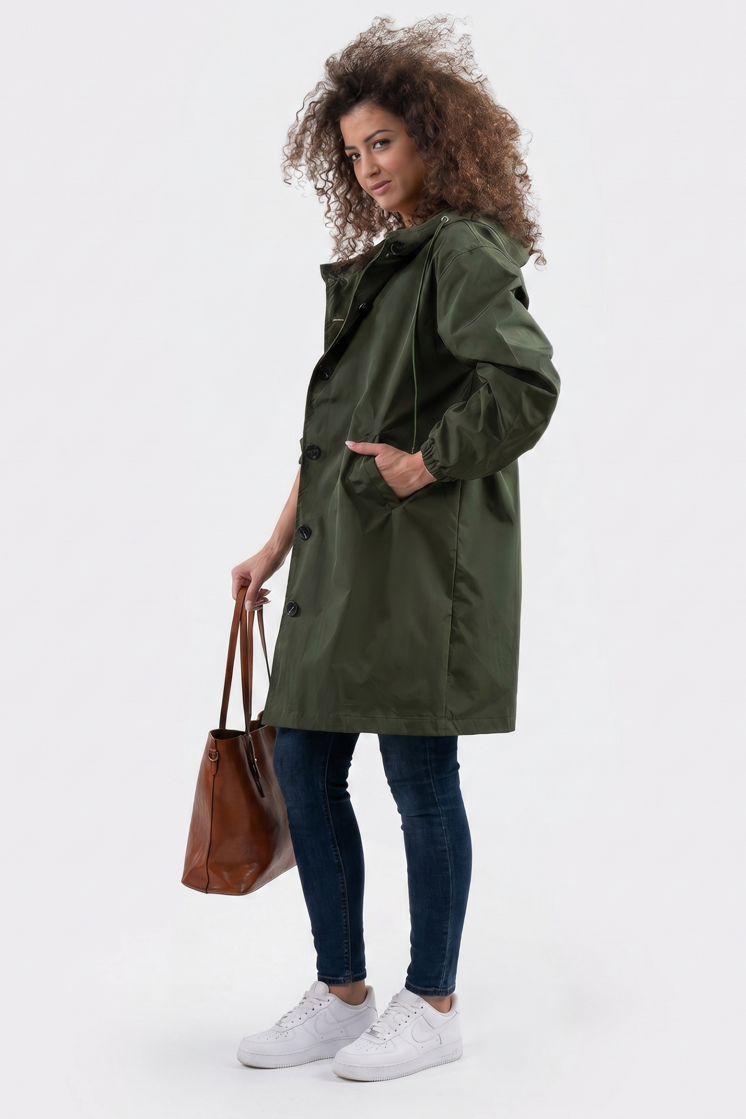 Wearing The Water Resistant Hooded Oversized Windbreaker Rain Jacket in olive green, blue jeans, and white sneakers, a person stands holding a brown leather tote bag against a plain white background.
