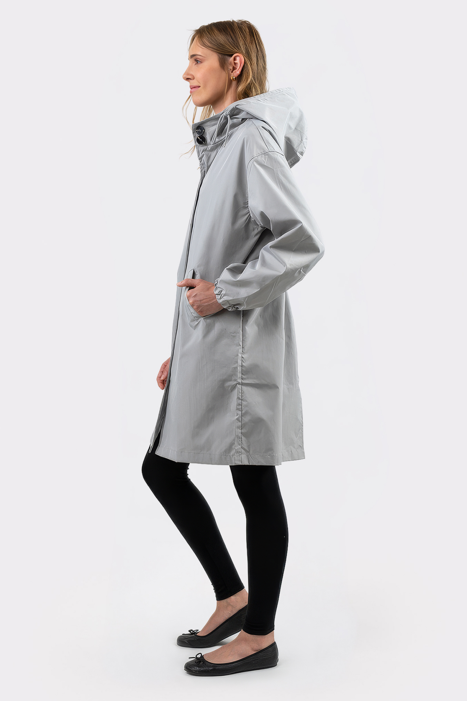 A person stands in profile against a plain white background wearing the Icon Windbreaker Rain Jacket in light gray, paired with black leggings and black flats, with their hands in the jacket’s pockets.