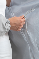 A person in a white outfit holds the light gray drawstring of the Icon Windbreaker Rain Jacket, tied in a knot, against sheer gray fabric. Only their torso and hand are visible.