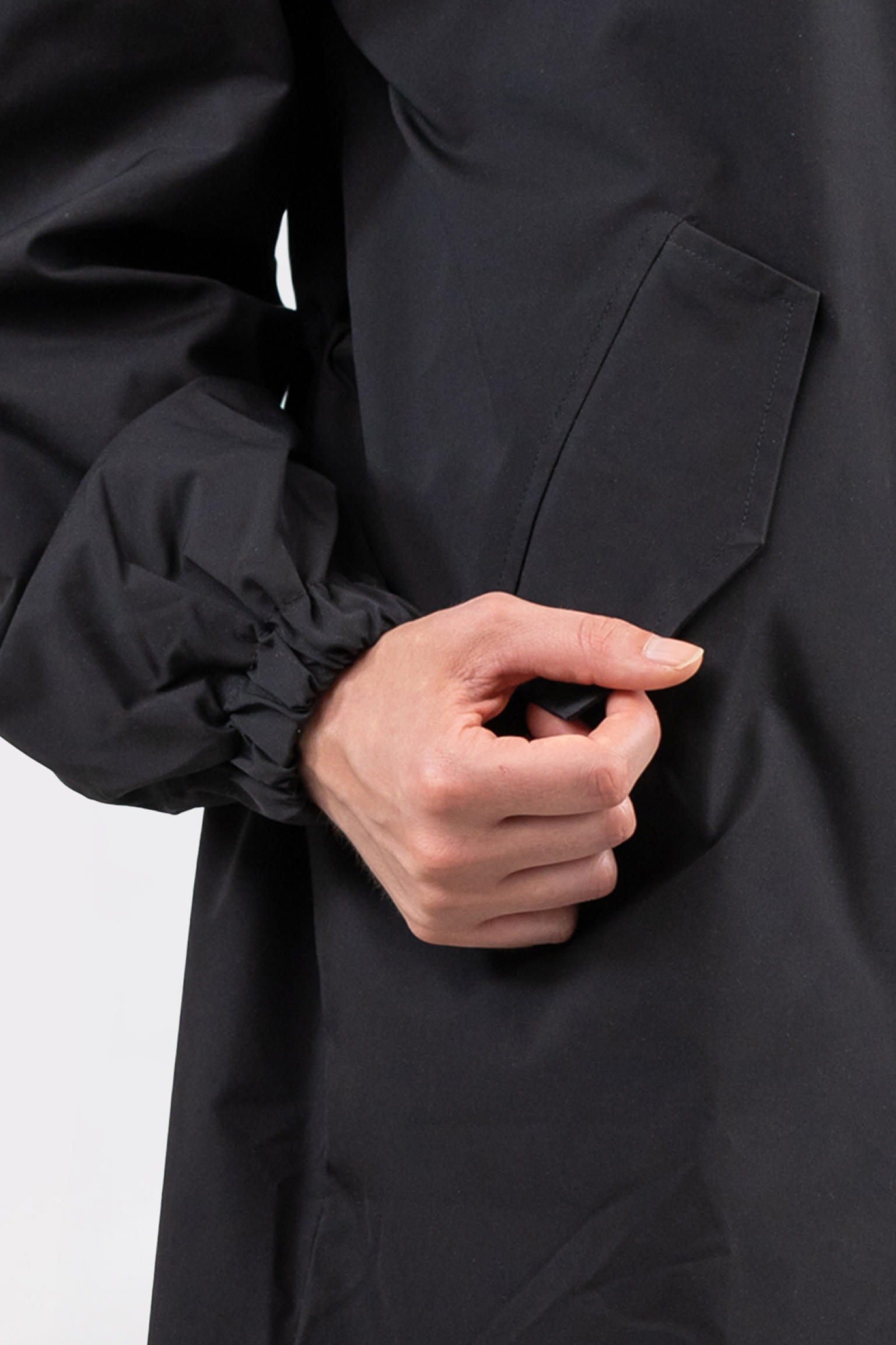 A person in The Water Resistant Hooded Oversized Windbreaker Rain Jacket, black and long-sleeved, holds the edge of its slanted pocket with their right hand against a plain light background.