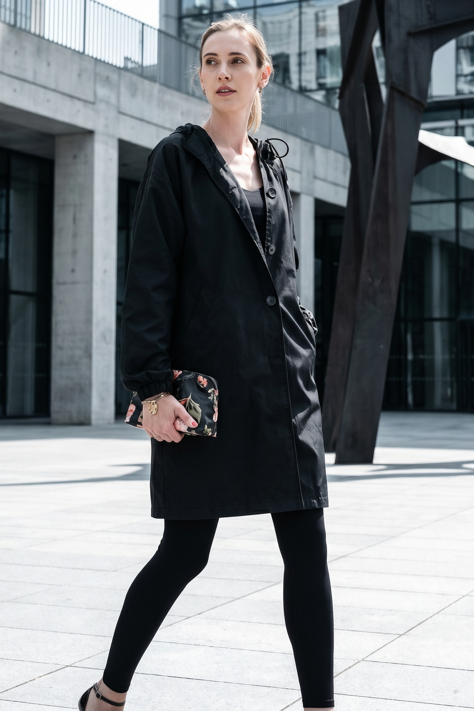 A woman wearing The Water Resistant Hooded Oversized Windbreaker Rain Jacket, black leggings, and black shoes walks outdoors in an urban setting. She holds a small floral-patterned clutch in front of modern concrete and glass buildings.