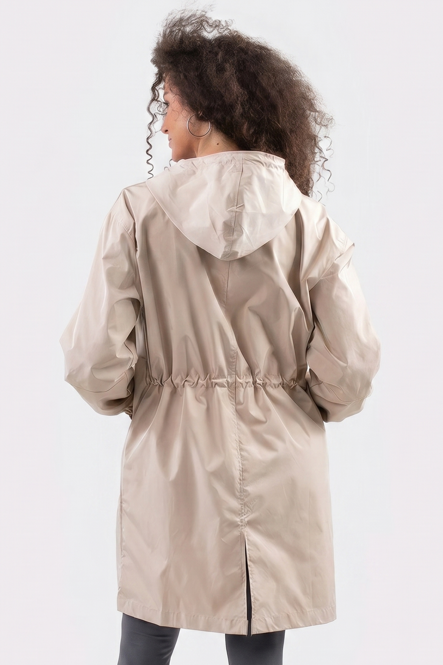 A person with curly hair is seen from the back wearing The Water Resistant Hooded Oversized Windbreaker Rain Jacket in light beige, featuring a gathered waist, center seam, and slit at the bottom, against a plain light background.