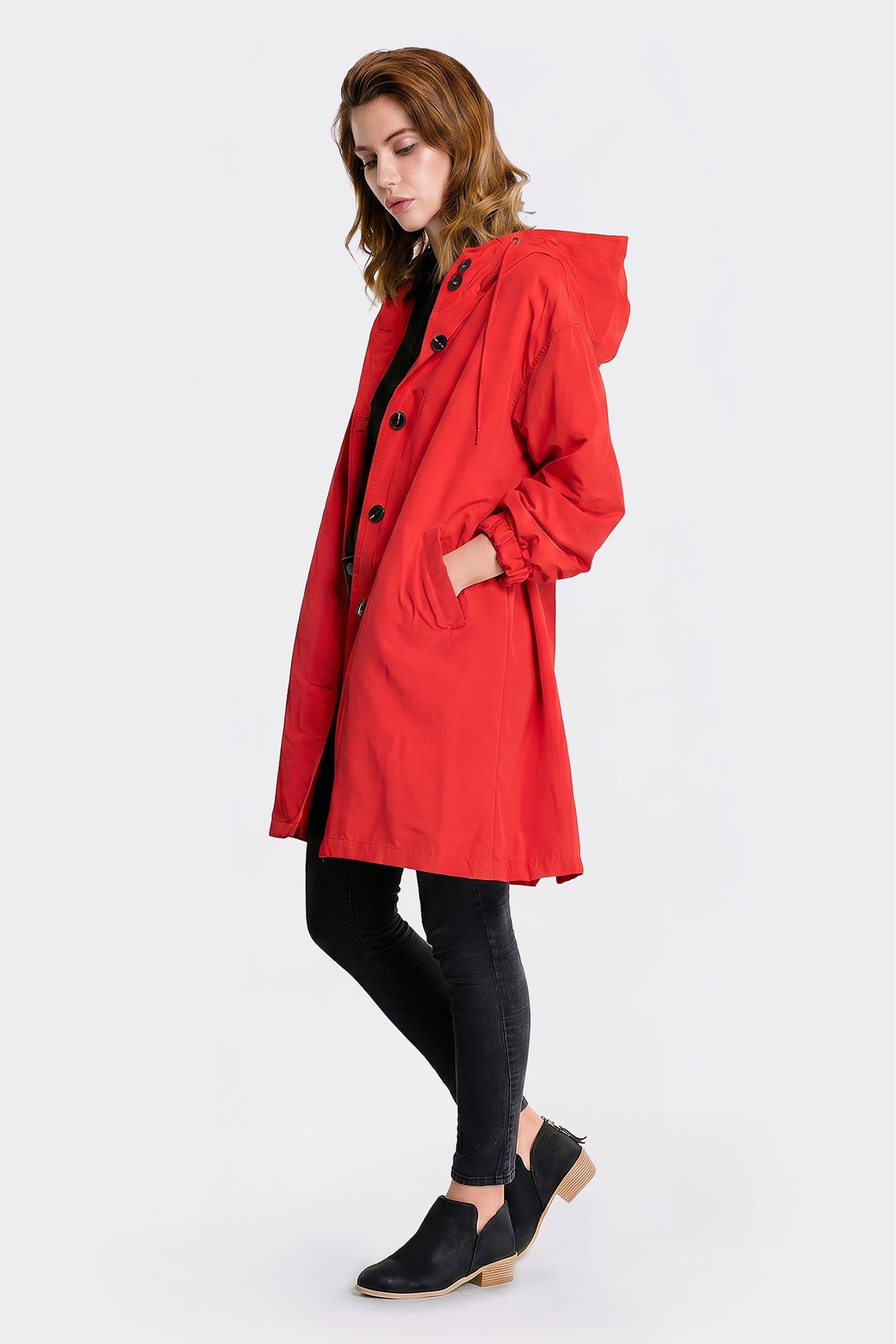 A person stands in profile wearing The Water Resistant Hooded Oversized Windbreaker Rain Jacket, which is long and red with large black buttons, paired with black pants and low-heeled black slip-on shoes. One hand rests in a jacket pocket; background is plain white.