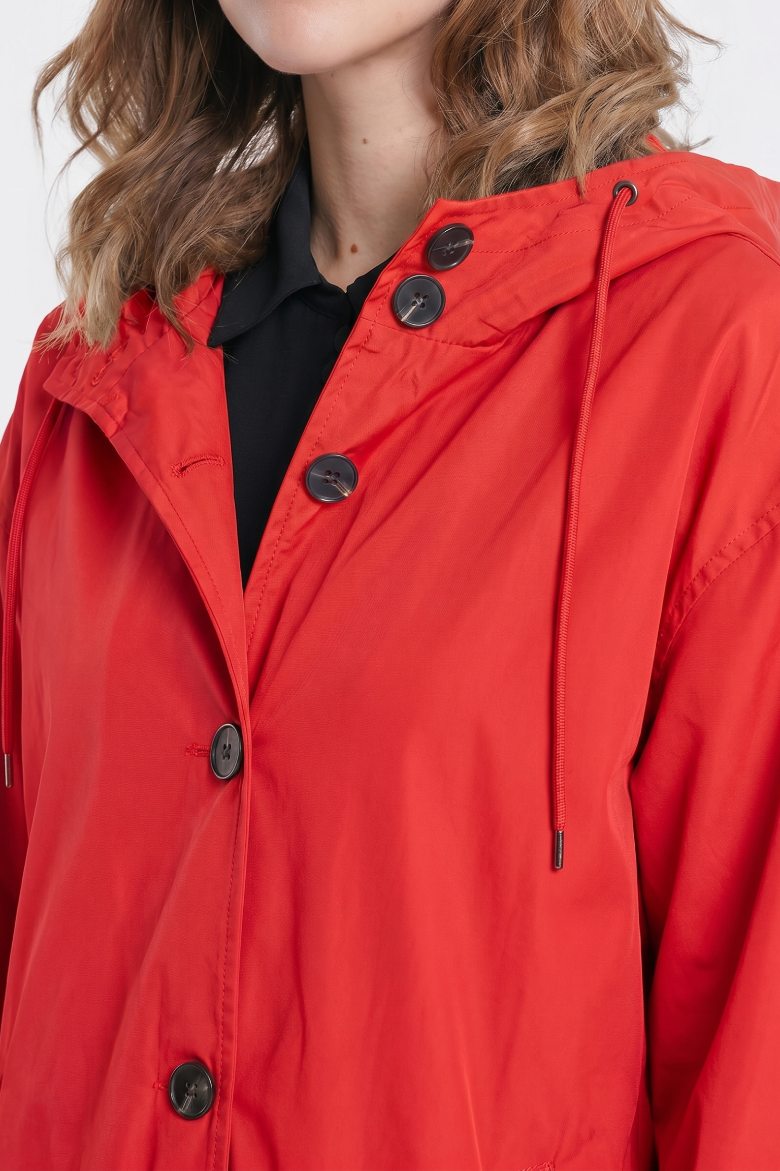 A person with wavy, light brown hair wears The Water Resistant Hooded Oversized Windbreaker Rain Jacket in bright red over a black top. Only the lower face, neck, and upper body are visible.