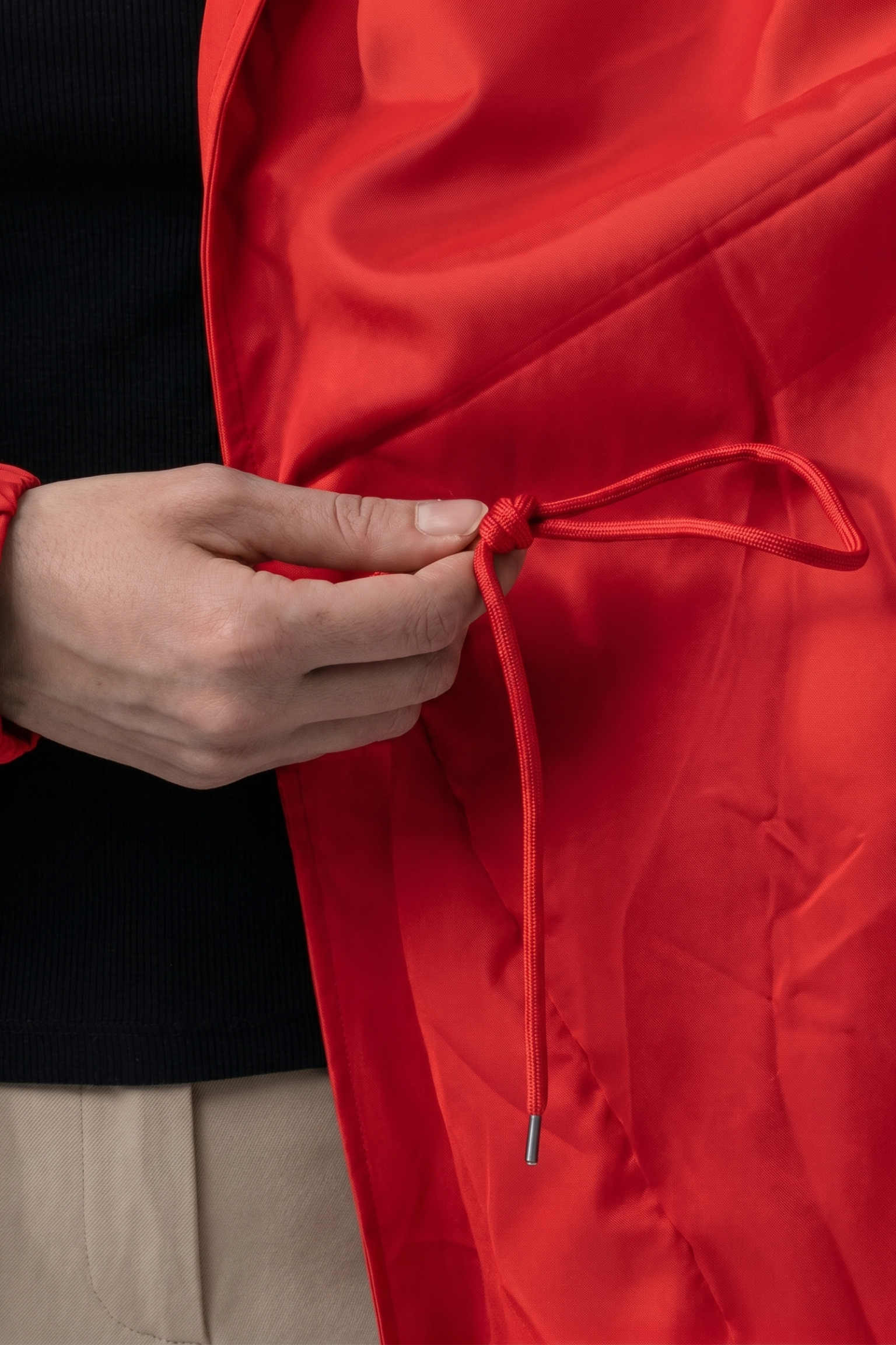 A person ties the drawstring inside the bright red Icon Windbreaker Rain Jacket while wearing beige pants and a black shirt. The photo highlights their hands and the jacket's interior.