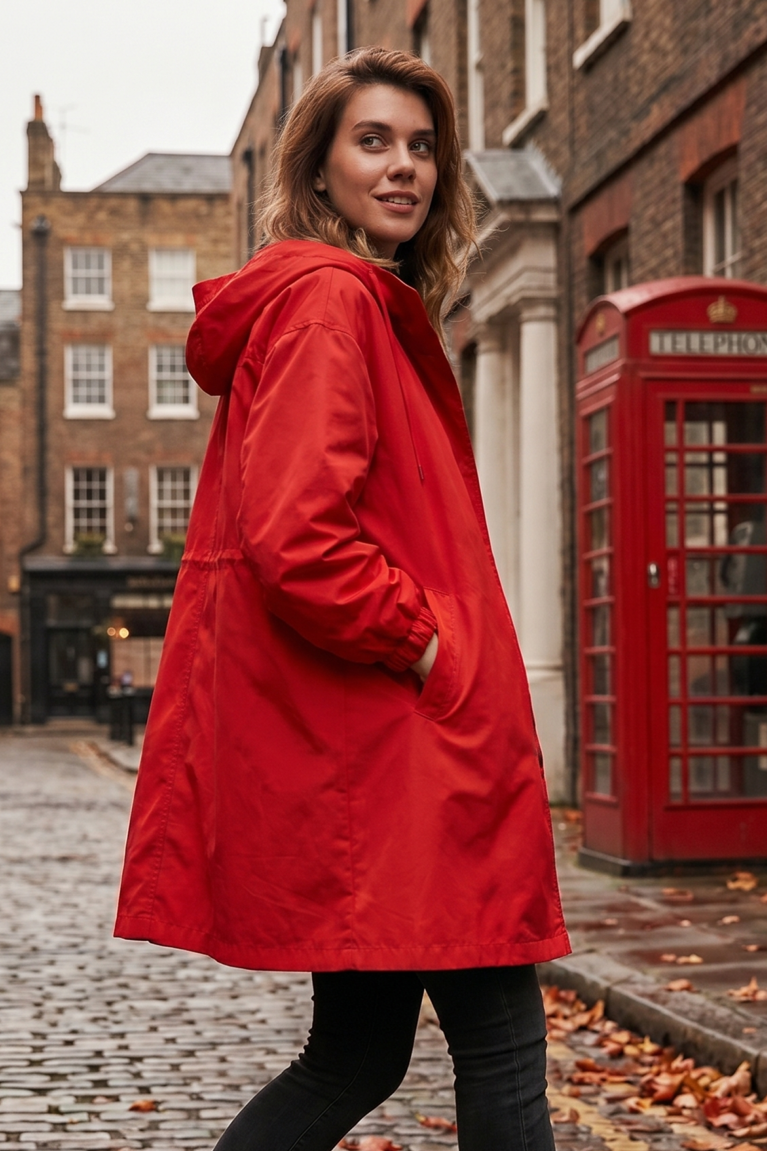 A person wearing The Water Resistant Hooded Oversized Windbreaker Rain Jacket in bright red stands on a cobblestone street beside a classic red British phone booth, with historic brick buildings in the background.