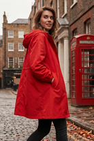 A woman wearing the Icon Windbreaker Rain Jacket stands on a cobblestone street beside a red British phone booth, with brick buildings and autumn leaves in the background.