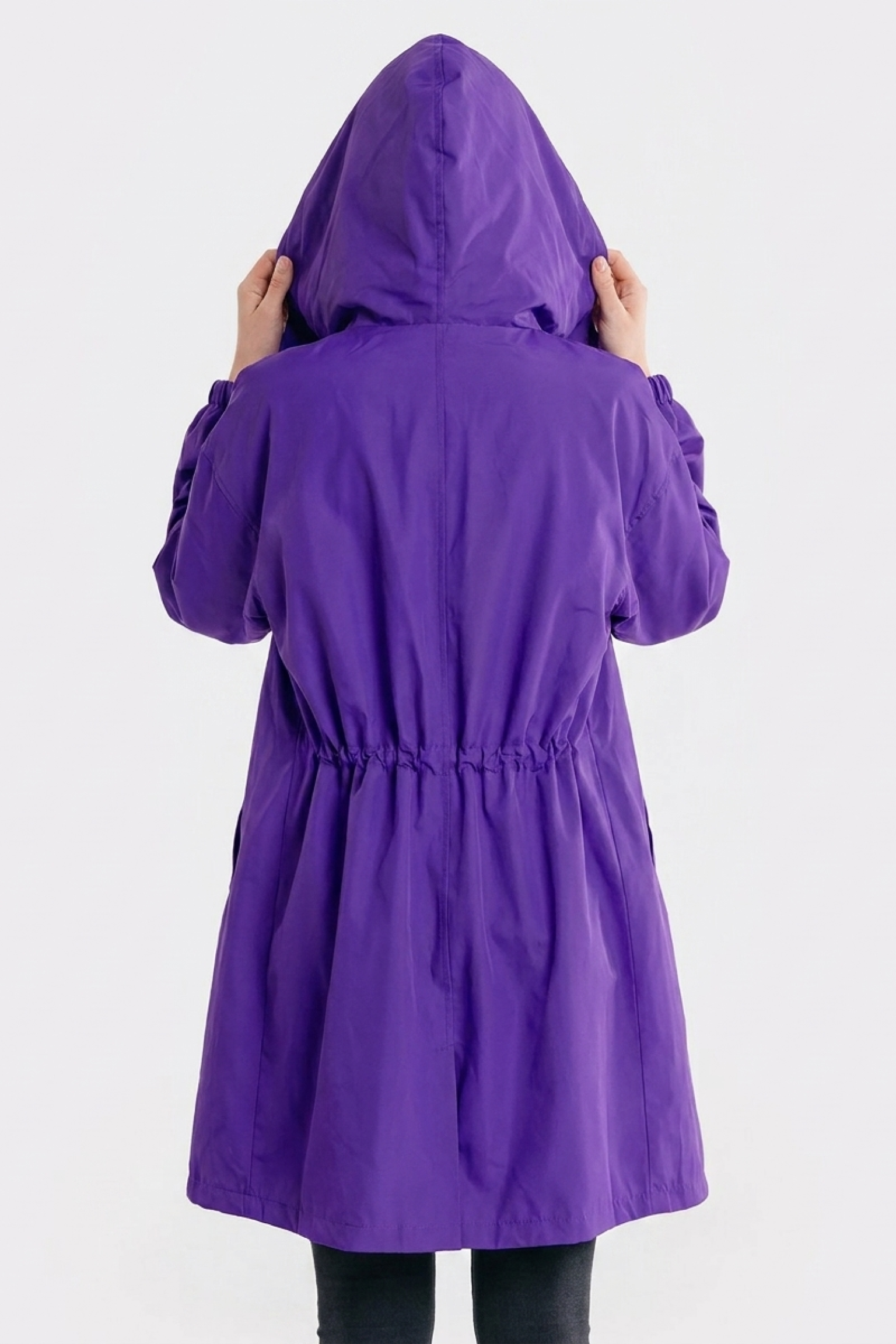 A person in The Water Resistant Hooded Oversized Windbreaker Rain Jacket, a long purple coat with a hood, stands with their back to the camera and holds the hood with both hands against a plain light background.
