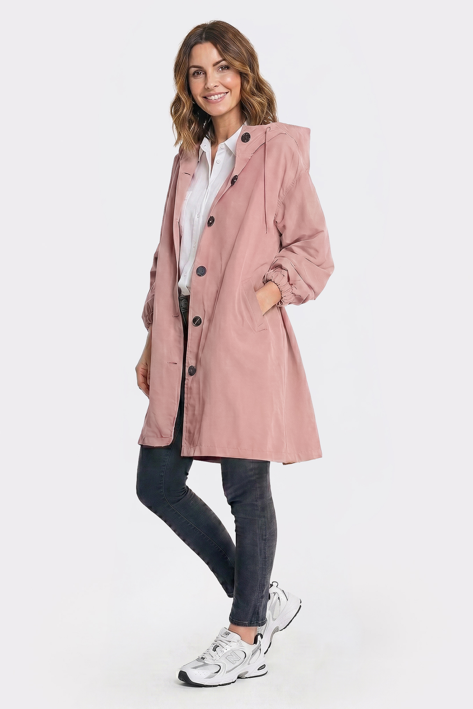 A woman with shoulder-length brown hair smiles at the camera, hands in pockets, wearing The Water Resistant Hooded Oversized Windbreaker Rain Jacket in light pink over a white shirt, paired with dark jeans and white sneakers against a plain background.