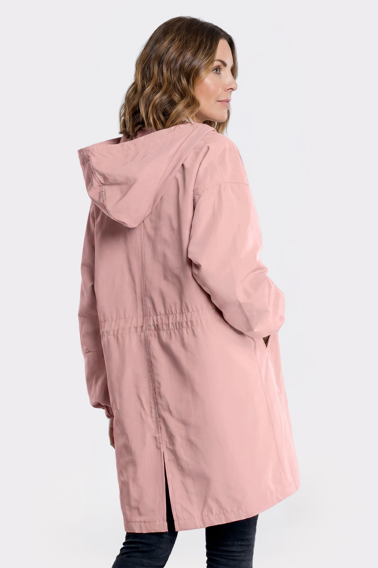 A woman with wavy brown hair is wearing The Water Resistant Hooded Oversized Windbreaker Rain Jacket in light pink. She stands against a plain white background, angled to show the back and side of the jacket.
