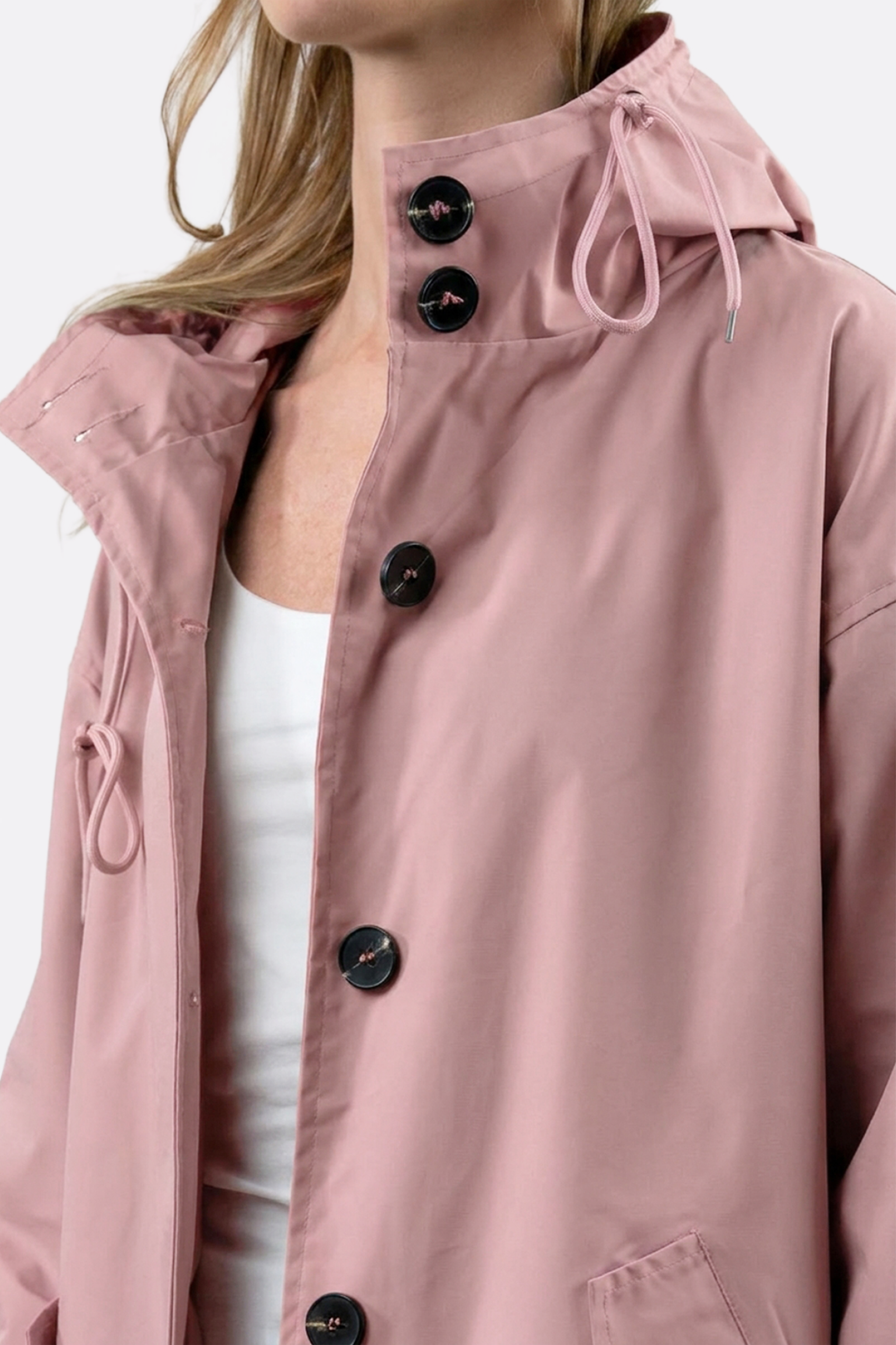 A person wearing The Water Resistant Hooded Oversized Windbreaker Rain Jacket in light pink, featuring black buttons and collar drawstrings, over a white top. Shown from shoulders to chest.