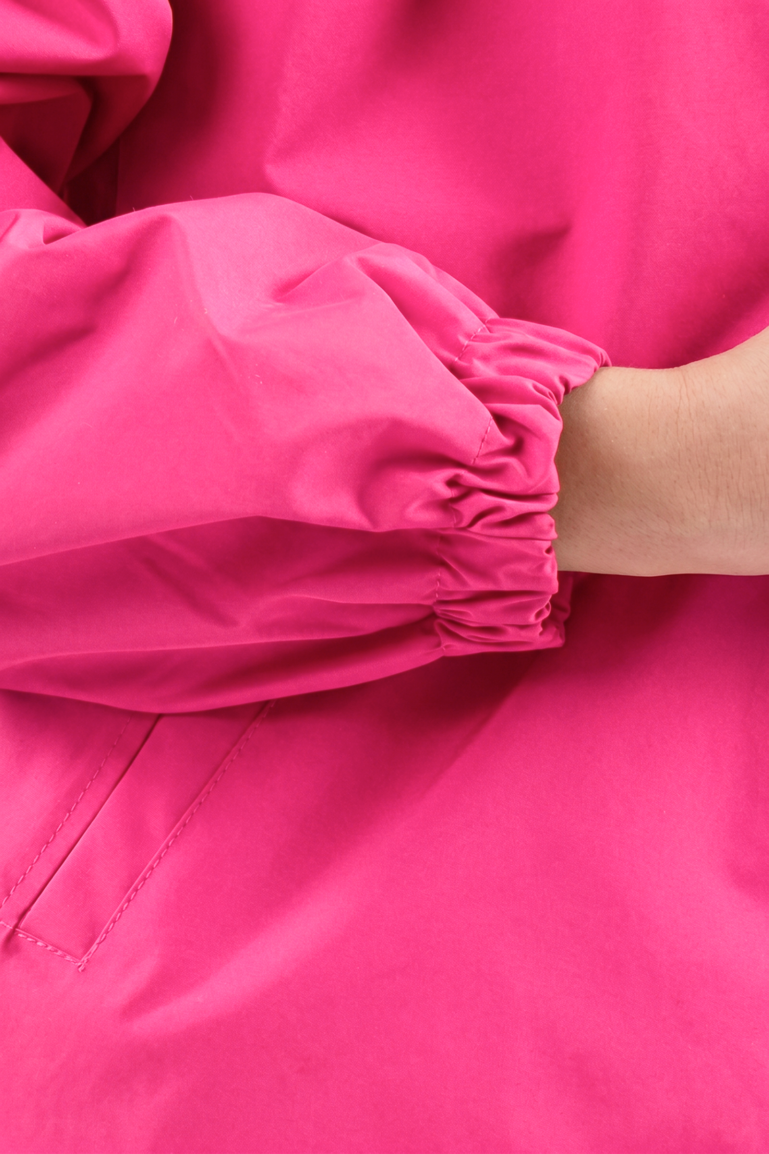 A close-up of a person’s arm wearing The Water Resistant Hooded Oversized Windbreaker Rain Jacket in bright pink shows its smooth, slightly shiny fabric, elastic cuff, and part of their hand.