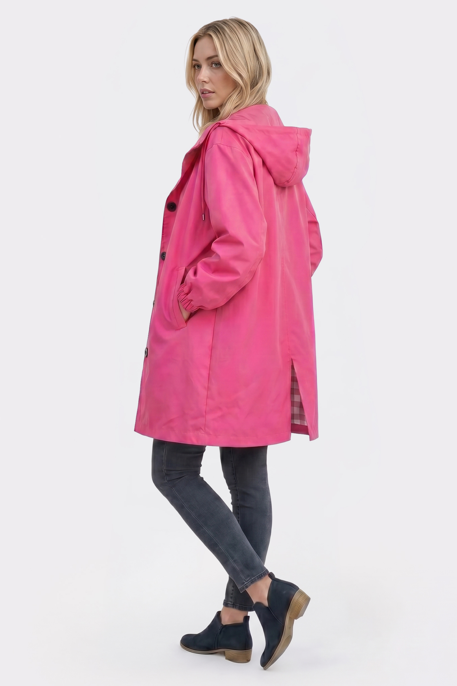 A person stands sideways in The Water Resistant Hooded Oversized Windbreaker Rain Jacket, which is bright pink with front buttons and side pockets. They pair it with dark skinny jeans and black ankle boots against a plain white background.