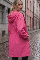 A person with blonde hair, wearing the bright pink Icon Windbreaker Rain Jacket, stands on a wet cobblestone street lined with old buildings and looks back over their shoulder.