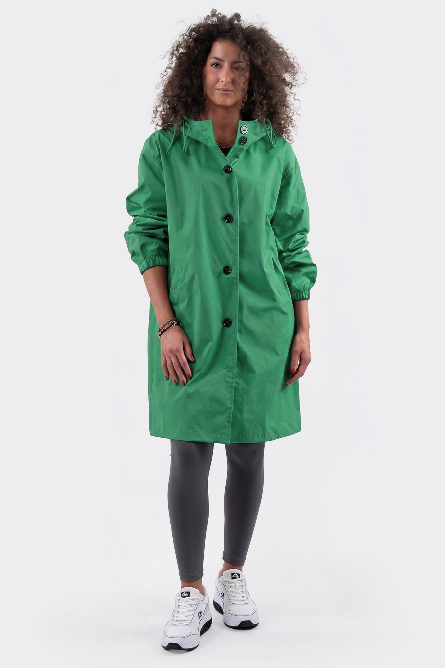 A person with curly hair wears The Water Resistant Hooded Oversized Windbreaker Rain Jacket in green, paired with dark leggings and white sneakers, standing against a plain white background.