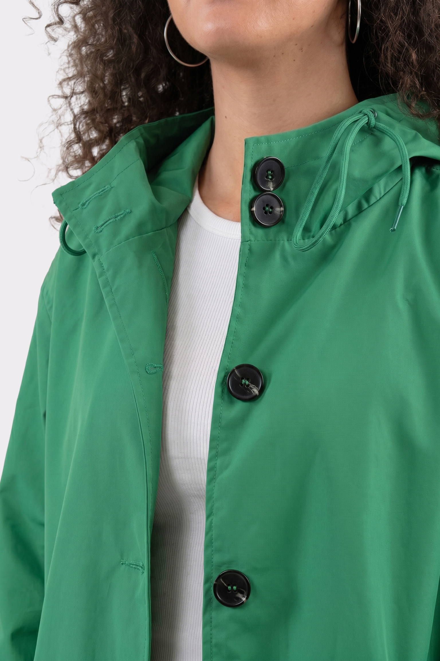 Wearing The Water Resistant Hooded Oversized Windbreaker Rain Jacket in green over a white ribbed shirt, the person’s lower face and upper chest are visible. They have curly hair and large hoop earrings. Black buttons accent the jacket.