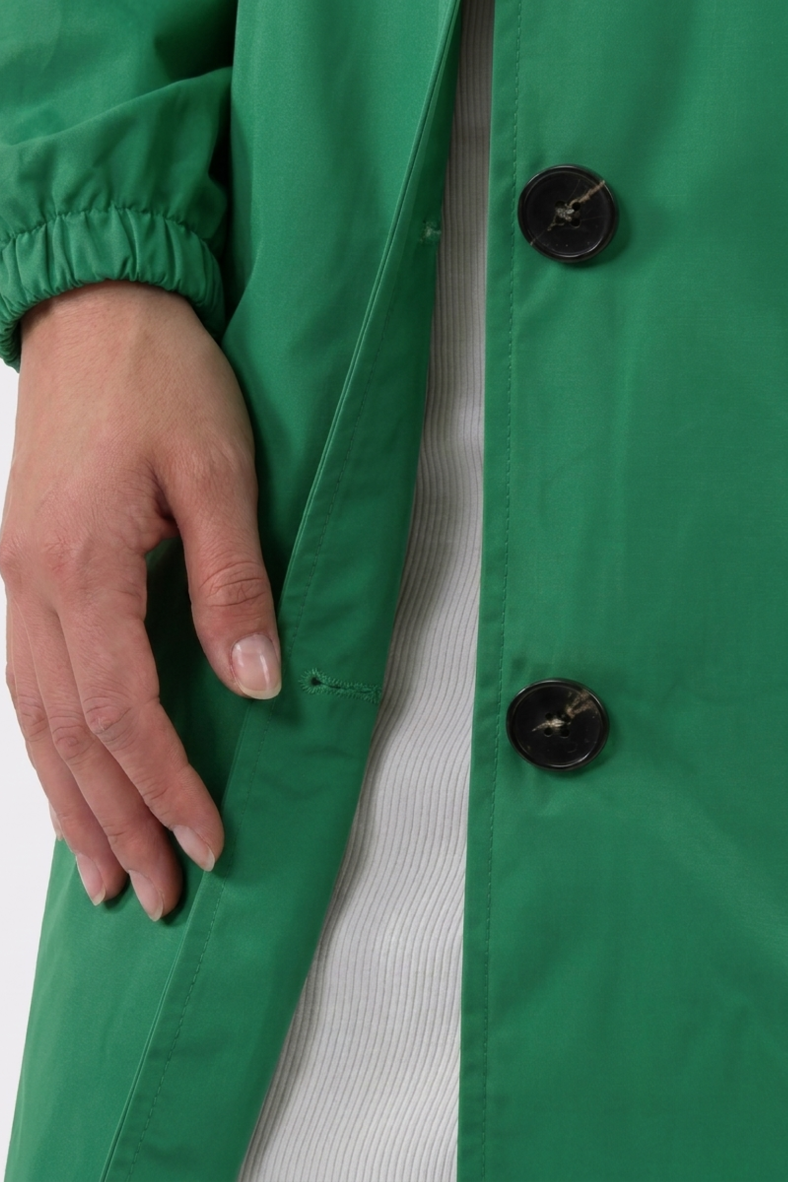 A person wearing The Water Resistant Hooded Oversized Windbreaker Rain Jacket has their left hand beside their body; the green coat with black buttons is open, showing a white ribbed garment underneath.