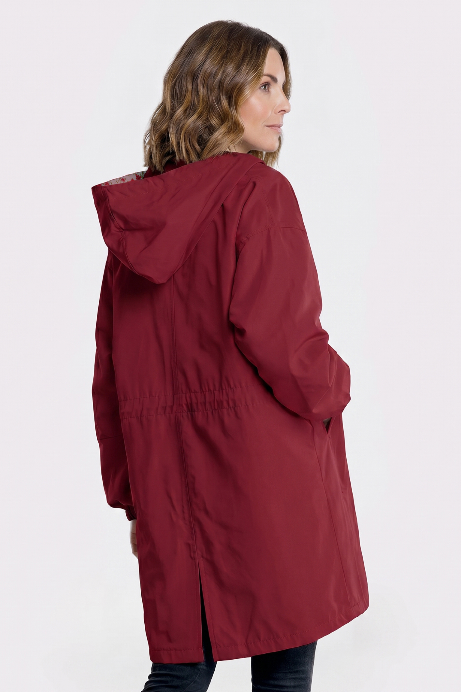 A person with shoulder-length wavy hair is seen from behind wearing the Icon Windbreaker Rain Jacket, a long, hooded red coat with a bottom slit and loose fit, standing against a plain light background.