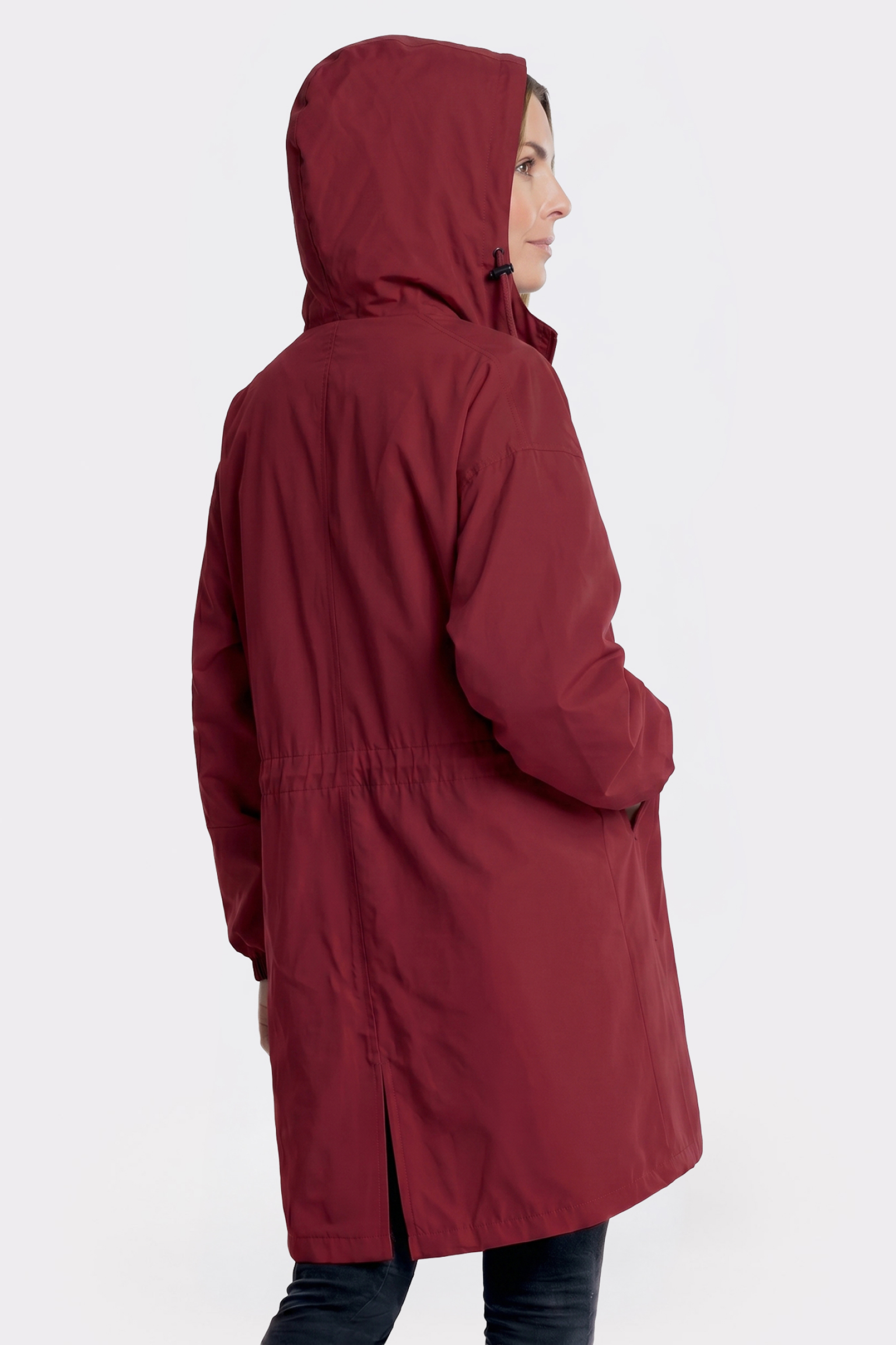 A person wears The Water Resistant Hooded Oversized Windbreaker Rain Jacket in red, featuring a long fit, drawstring waist, back slit, and hood. They stand sideways with one hand in a pocket against a plain white background.