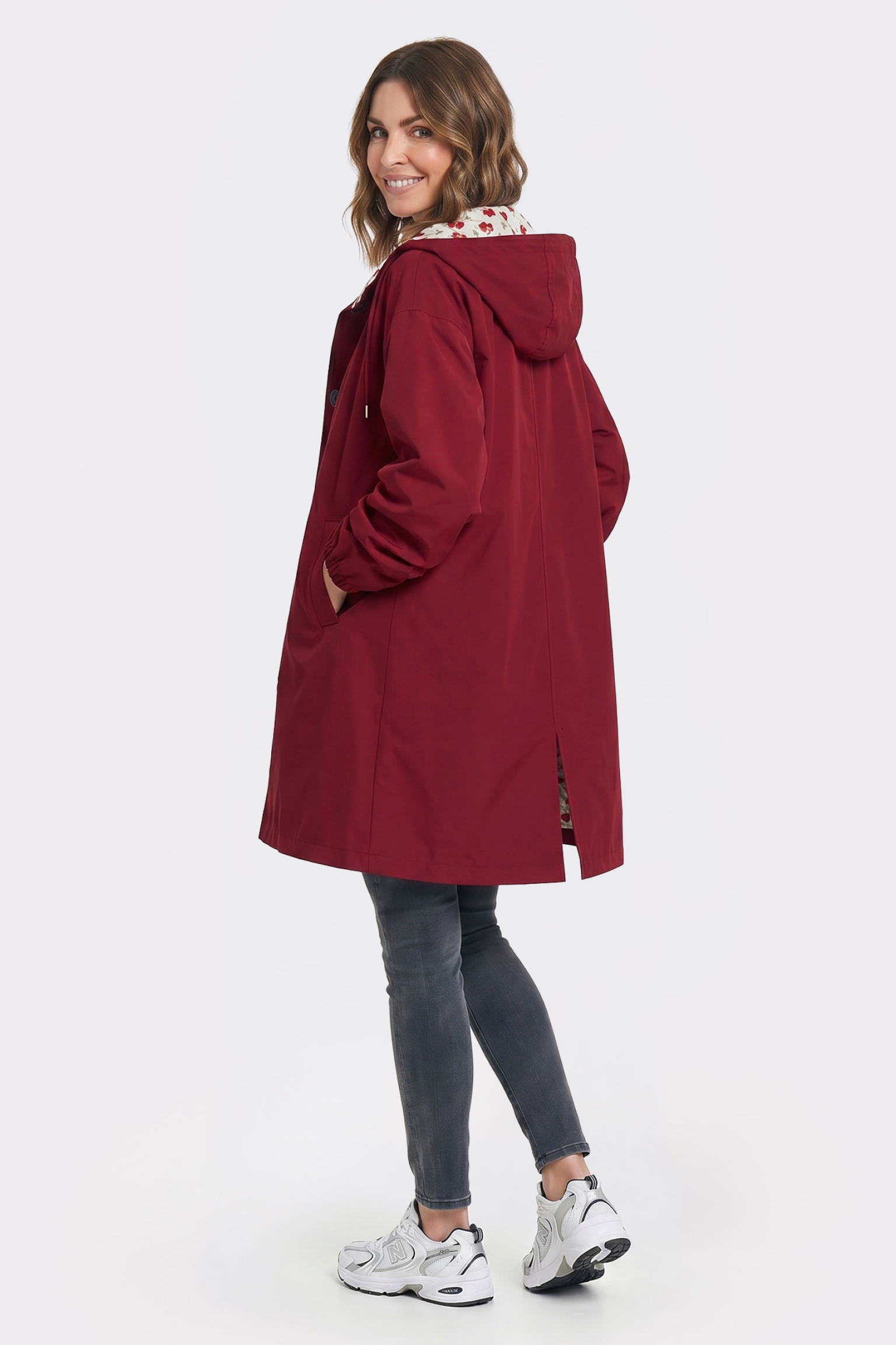 A person with shoulder-length wavy hair wears The Water Resistant Hooded Oversized Windbreaker Rain Jacket in maroon, paired with gray jeans and white sneakers, standing and looking over their shoulder against a plain white background.