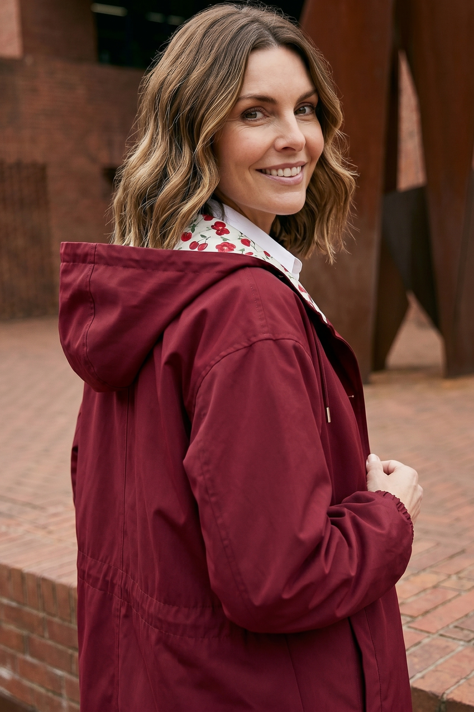 A woman with wavy brown hair smiles over her shoulder, wearing The Water Resistant Hooded Oversized Windbreaker Rain Jacket in maroon and a white floral shirt. Brick walls and abstract metal sculptures form the background.
