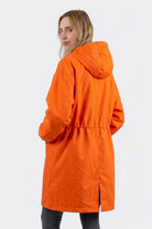 A person with long blonde hair wears the Icon Windbreaker Rain Jacket in orange and black pants, standing against a plain white background with their back to the camera and looking over their shoulder.