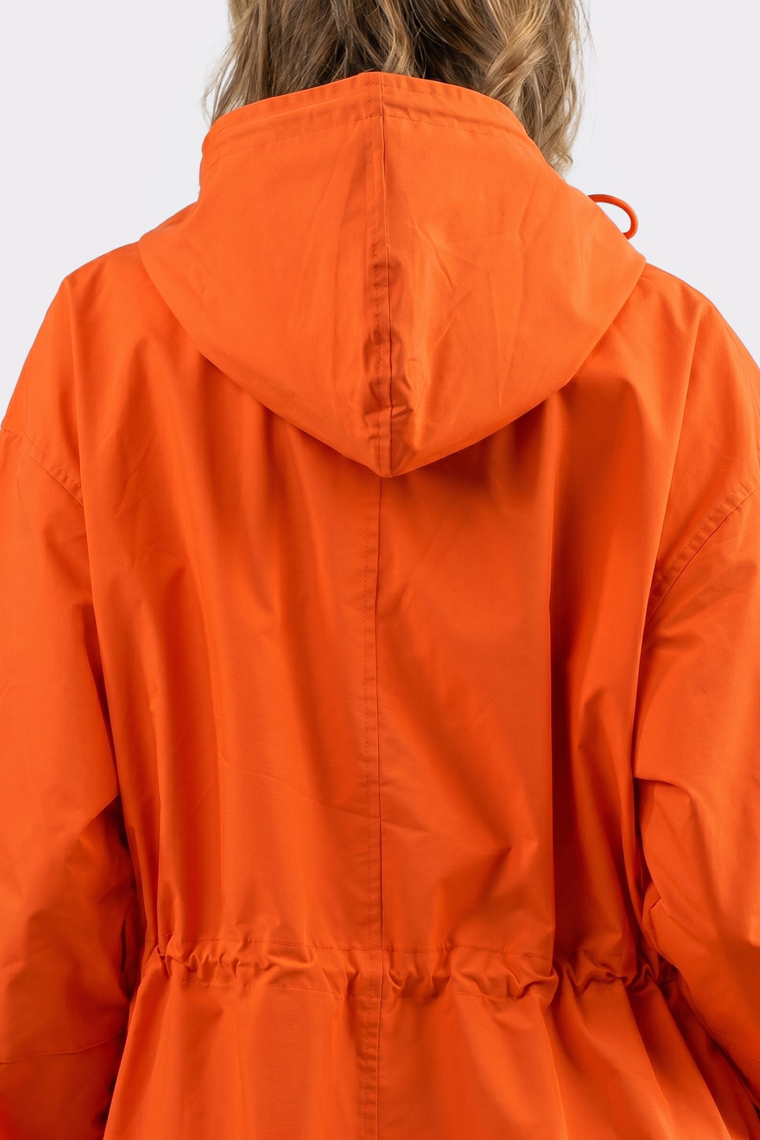 A person is shown from the back wearing The Water Resistant Hooded Oversized Windbreaker Rain Jacket in orange, featuring a loose fit with a cinched waist. Their wavy, light brown hair is partially visible.