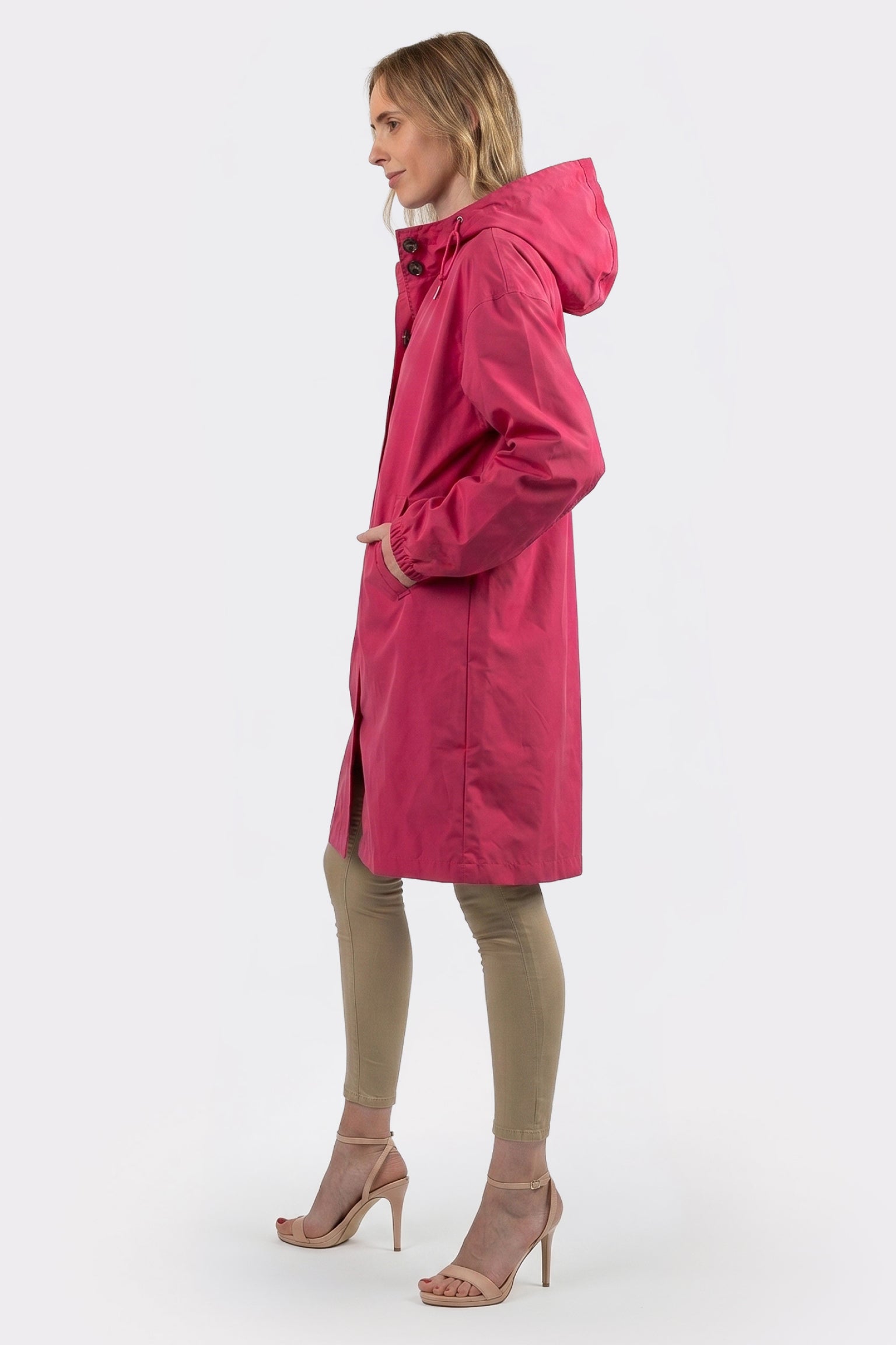 A woman stands in profile wearing the Icon Windbreaker Rain Jacket, beige fitted pants, and light pink high-heeled sandals against a plain white background.