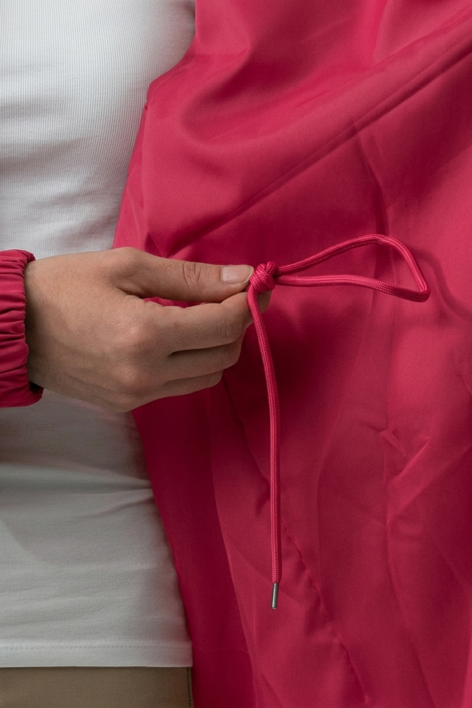 A person in a white shirt adjusts the pink drawstring, tied in a bow, on The Water Resistant Hooded Oversized Windbreaker Rain Jacket’s pink fabric.